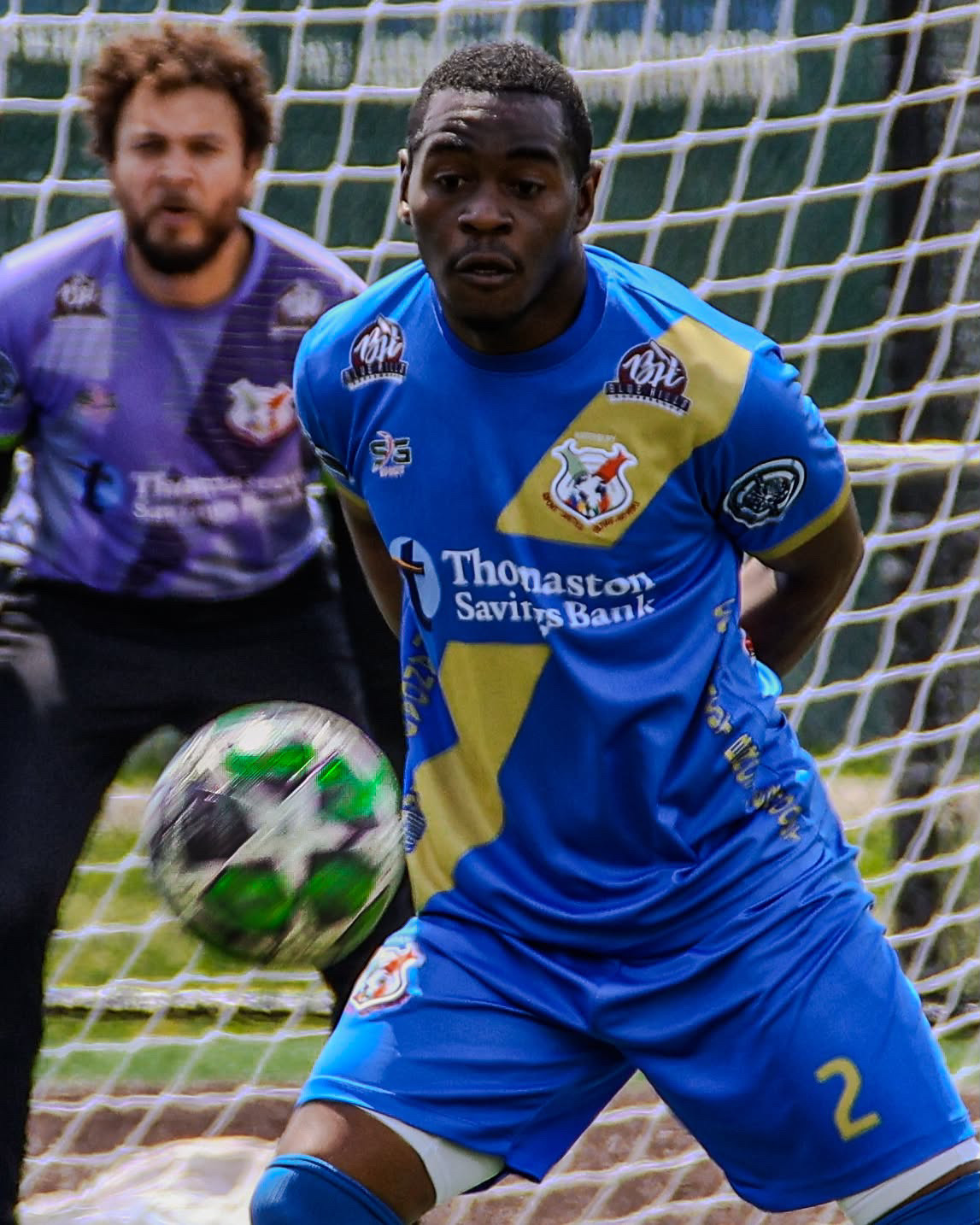 Defender in blue and gold tracking a spinning ball with the keeper set behind him in the frame.