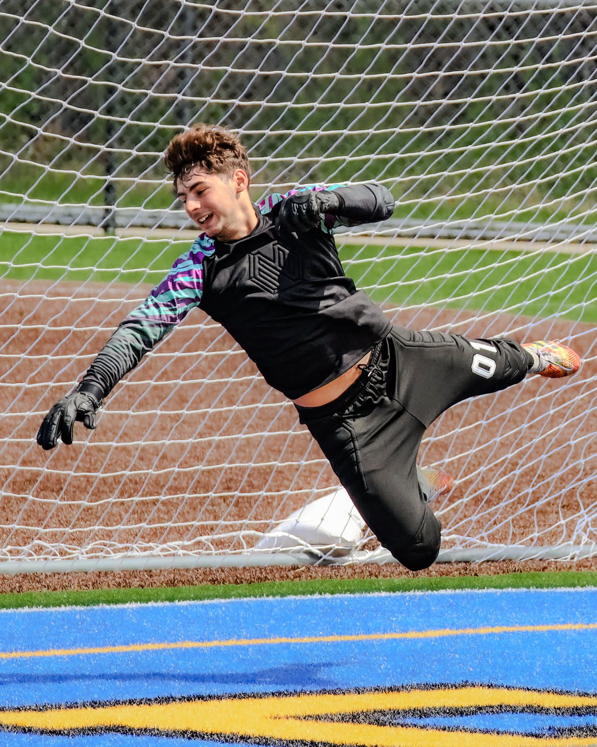 Young keeper in black stretched fully airborne inside the six-yard box, gloves reaching for the ball.