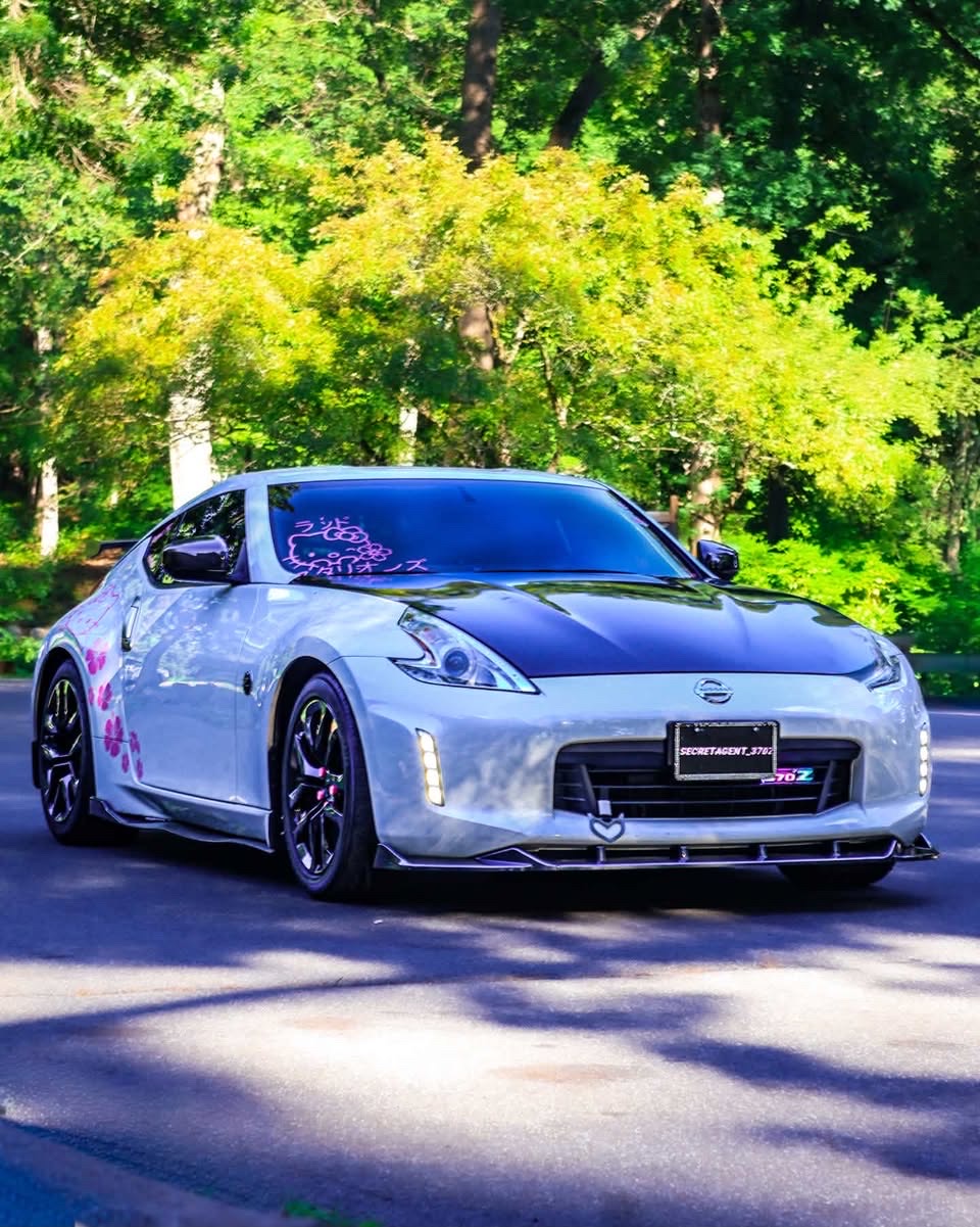 Front-quarter view of the 370Z on a shaded forest road, headlights on and cherry-blossom decals catching light.