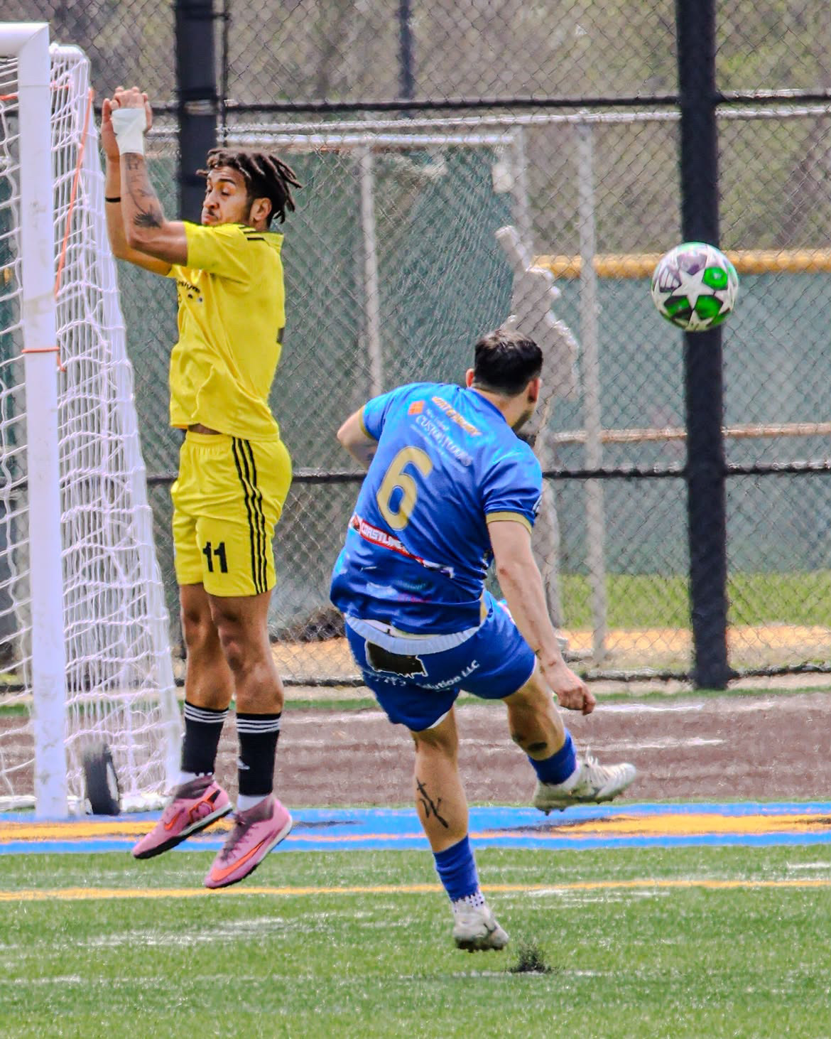 Goalkeeper in yellow rising to punch clear as a striker in blue follows through on a shot.